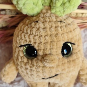 May include: A handmade, plush toy in the shape of a root vegetable with a smiling face. The toy is beige with green leaves on top and large, glittery green eyes. The plushie is made of soft, textured yarn.