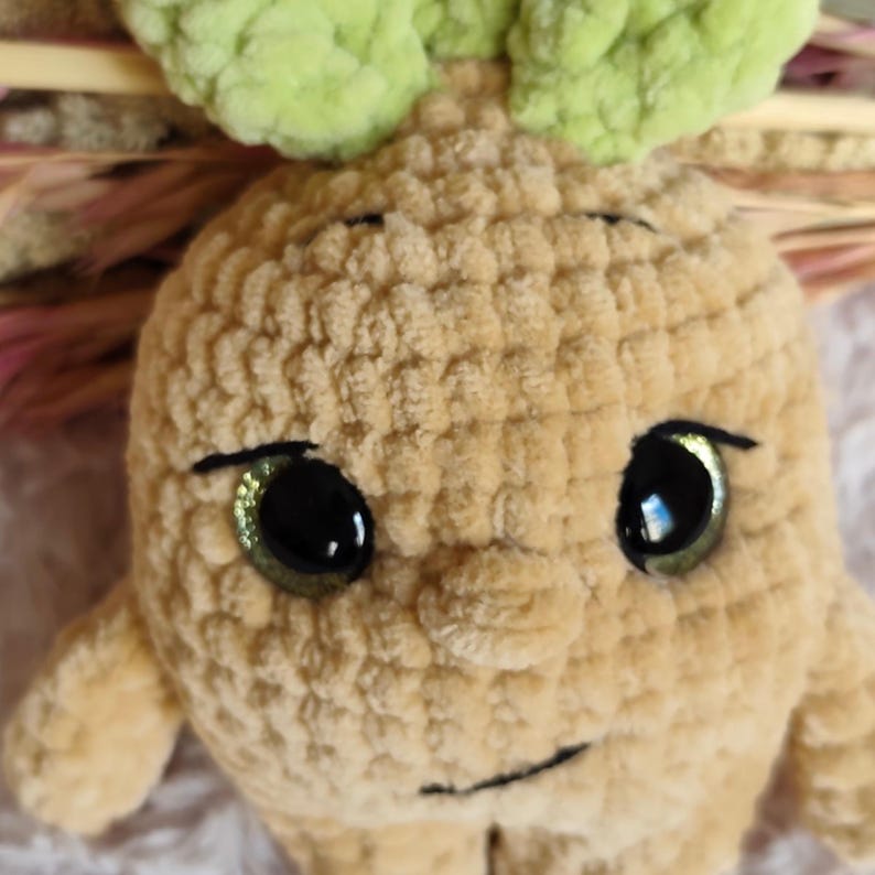 May include: A handmade, plush toy in the shape of a root vegetable with a smiling face. The toy is beige with green leaves on top and large, glittery green eyes. The plushie is made of soft, textured yarn.