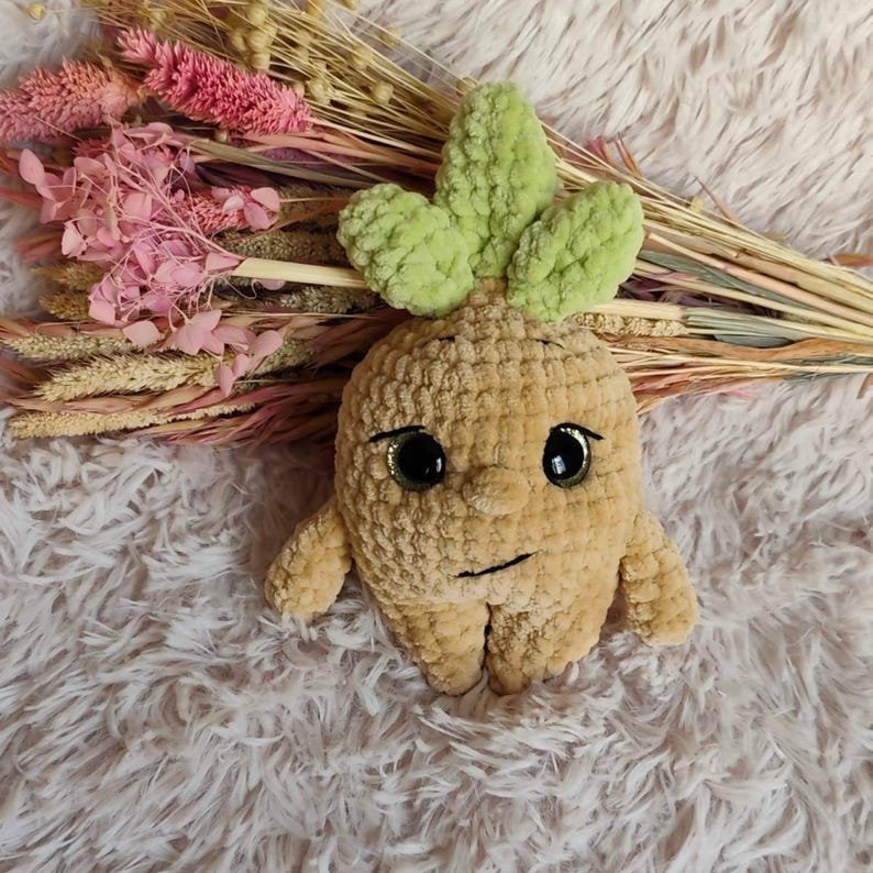 May include: A handmade, tan-colored crochet doll with a smiling face, large black eyes, and green leaves on top. The doll has small arms and legs. Dried pink and beige flowers are in the background.