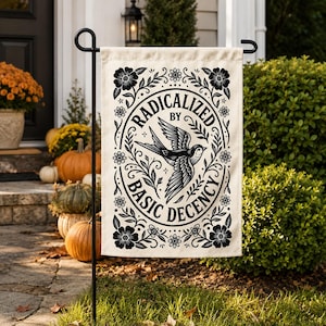 May include: A rectangular cream-colored garden flag featuring a black ink-style illustration of a bird in flight at the center, surrounded by a floral border and the text RADICALIZED BY BASIC DECENCY printed in a bold, circular black font.