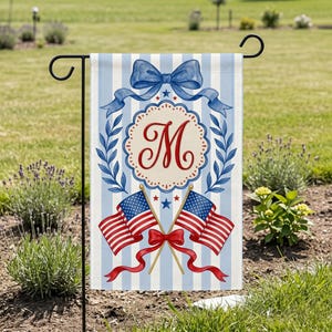 May include: A rectangular garden flag featuring a light blue and white vertical striped background. A large blue bow sits at the top above a central cream-colored scalloped circle containing a red letter M. Two crossed American flags are below.