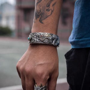 May include: A wide silver cuff bracelet featuring an intricate embossed dragon design with gold-toned accents and a small round turquoise stone inlay. The bracelet is worn on a wrist, accompanied by a matching silver ring with a textured surface.