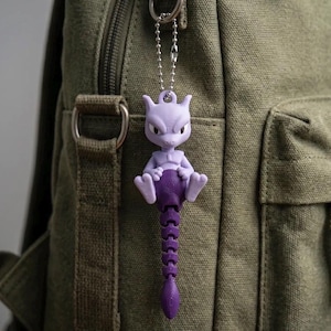 May include: A light purple and dark purple articulated Mewtwo character keychain featuring a ball chain attachment. The figure has a smooth, matte finish with a segmented, flexible tail extending downward from the seated character's body.