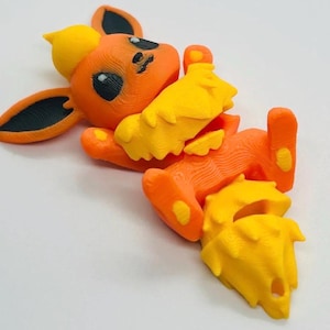 May include: A 3D printed articulated figurine of a fox-like creature with orange fur, a yellow chest ruff, and a yellow bushy tail. The creature features large pointed ears with black inner details, black eyes, and a small yellow tuft on its head.