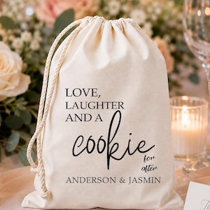 May include: A cream-colored fabric drawstring pouch featuring black printed text that reads: LOVE, LAUGHTER AND A cookie for after ANDERSON & JASMIN. The bag has a thick, braided cord closure and is made of a woven textile material.