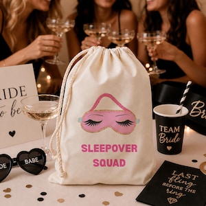 Peut inclure: Un sac en coton crème avec un motif de masque de sommeil rose et l'inscription "SLEEPOVER SQUAD" en rose. On y trouve également des verres à champagne, des lunettes de soleil en forme de cœur et une tasse "Team Bride".