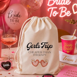 Może przedstawiać: Kremowa torba ze sznurkiem z napisem "Girls Trip CHEAPER THAN THERAPY" w kolorze czarnym i grafiką różowych okularów przeciwsłonecznych. Różowe balony i neonowy znak z napisem "Bride To Be" w tle.