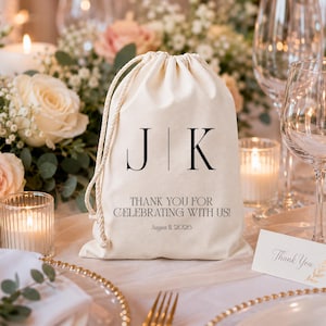 Peut inclure: Un sac en coton crème avec cordon de serrage, orné des initiales "J K" en noir et du texte "THANK YOU FOR CELEBRATING WITH US! August 11, 2026." Le sac est posé sur une table avec des compositions florales, des bougies et des couverts.
