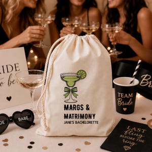 May include: A cream drawstring bag featuring a margarita design and the text "MARGS & MATRIMONY JANE'S BACHELORETTE". Also present are a "Team Bride" cup, heart-shaped sunglasses, and a black napkin.
