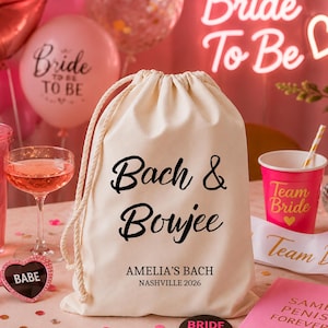 May include: A cream-colored drawstring bag with the words "Bach & Boujee" in black script. Below, it reads "AMELIA'S BACH NASHVILLE 2026." The background includes pink balloons, a neon sign, and a pink cup that says "Team Bride."