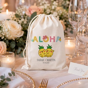 May include: A cream-colored drawstring bag with the word "ALOHA" in colorful letters and two cartoon pineapples. The bag also includes the names "Sarah + Martin" and the date "10.04.2026". The bag is on a table with floral arrangements and candles.