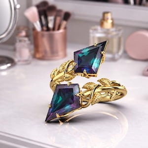 May include: A gold ring featuring two kite-shaped, color-shifting teal and purple gemstones. The band is designed with a detailed vine and leaf motif that wraps around the finger, holding the two stones in a bypass style setting.