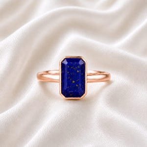 Könnte beinhalten: Ein roségoldener Ring mit einem rechteckigen, tiefblauen Edelstein mit Goldsprenkeln. Der Edelstein ist in einer Fassung gefasst. Der Ring wird auf weißem, seidigem Stoff präsentiert.