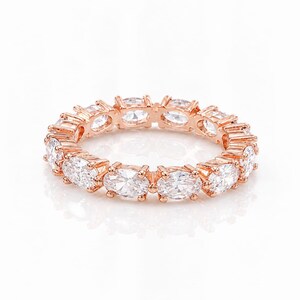 Könnte beinhalten: Ein Eternity-Ring aus Roségold mit einer durchgehenden Reihe funkelnder, oval geschliffener, klarer Steine, die einzeln in Krappen gefasst sind. Das Design zeigt eine gleichmäßige Anordnung facettierter Edelsteine rund um den gesamten Ring.
