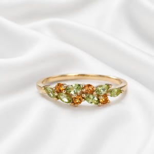 Könnte beinhalten: Ein goldener Ring mit grünen und orangen Edelsteinen. Die Edelsteine sind in einem blattartigen Muster angeordnet. Der Ring ist vor einem weißen Hintergrund abgebildet.