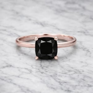 Könnte beinhalten: Roségoldener Ring mit einem großen, quadratisch geschliffenen schwarzen Edelstein. Der Edelstein wird von vier Krappen gehalten. Das Ringband ist schlicht und poliert. Der Ring ist vor einem Marmorhintergrund platziert.