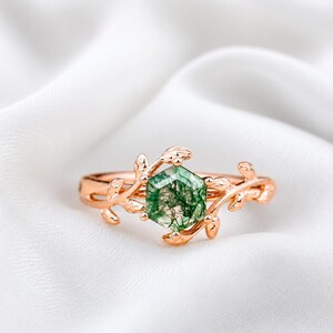 Könnte beinhalten: Ein roségoldener Ring mit einem sechseckigen grünen Moosachat-Edelstein. Das Ringband ist mit filigranen Blatt- und Rankendetails verziert, die ein von der Natur inspiriertes Design schaffen. Der Ring ist vor einem weißen Hintergrund platziert.