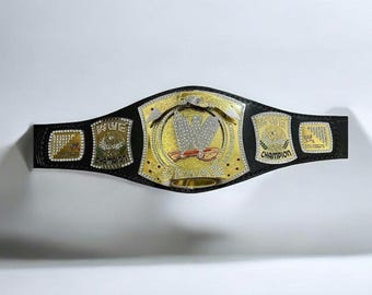 May include: A black leather championship belt featuring a large central gold plate with a silver eagle emblem and the letters WWE encrusted with clear stones. The belt includes four smaller gold side plates with engraved text and stone accents.