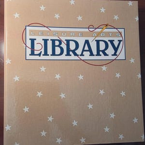 May include: A tan hardcover book with a repeating pattern of small white stars across the cover. The center features a rectangular logo with the text LEISURE ARTS LIBRARY in dark blue, accented by a red thread graphic and a gold sewing needle.