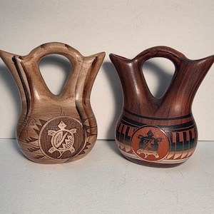 May include: Two ceramic vessels with arched handles and flared rims. The left vessel is light brown with dark brown wood-grain patterns and a turtle motif. The right vessel is dark reddish-brown with black and orange geometric accents and a turtle design.