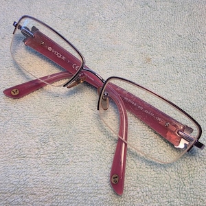 May include: A pair of Vogue eyeglasses with a pink frame and clear lenses. The glasses have a semi-rimless design with a metal bridge and temples. The Vogue logo is visible on the frame.