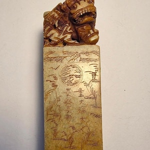 May include: A rectangular beige stone seal featuring a carved lion figure on top. The front surface displays intricate line engravings of landscape elements, a circular emblem, and Chinese characters including 量大福大 and 長樂長壽.