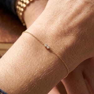 May include: A delicate gold chain bracelet featuring two small, round, clear stones set side-by-side in the centre. The bracelet is worn on the wrist, accompanied by a gold metal watch with a link band visible on the same arm.