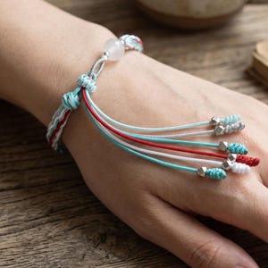 May include: A woven cord bracelet featuring a light pink spherical bead, multiple dangling strands in shades of turquoise, white, and red, and small metallic silver-toned beads at the ends of each cord, secured with a sliding knot closure.
