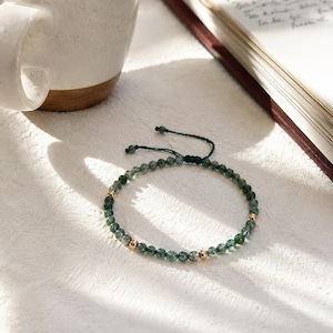 May include: A green beaded bracelet with gold-colored accents. The bracelet is made of small, round, green beads and features three gold-colored beads. The bracelet is on a white surface, next to a white mug and an open book.