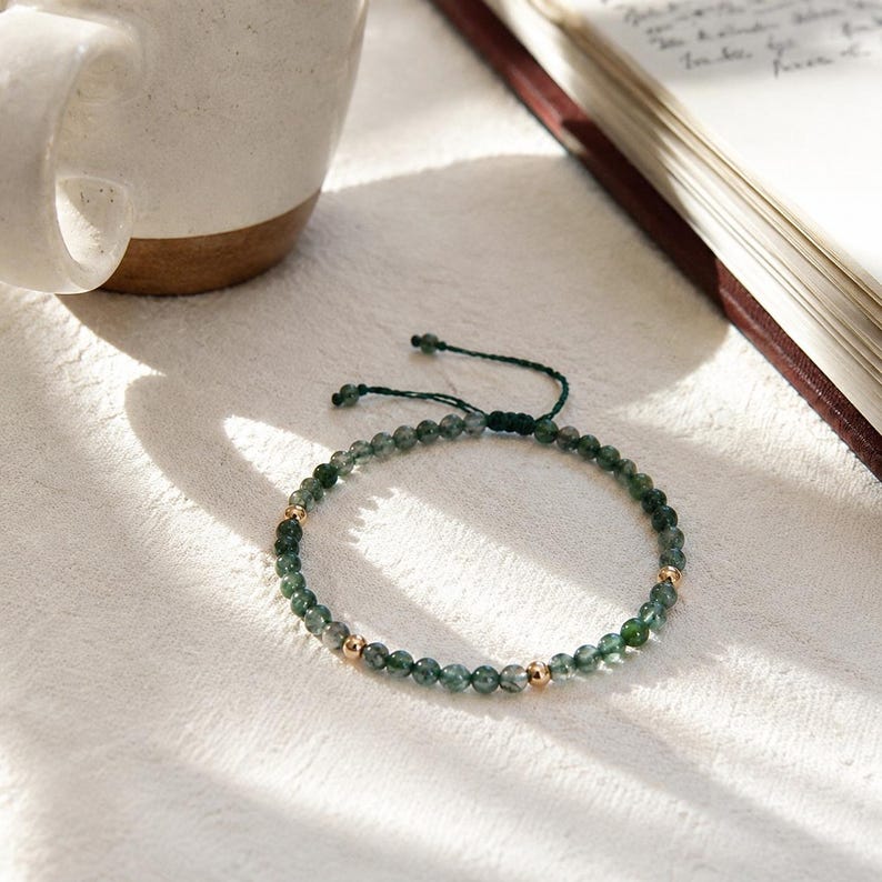 May include: A green beaded bracelet with gold-colored accents. The bracelet is made of small, round, green beads and features three gold-colored beads. The bracelet is on a white surface, next to a white mug and an open book.