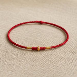 Puede incluir: Una pulsera fina de cord&oacute;n rojo con una cuenta central dorada mate, flanqueada por dos peque&ntilde;as envolturas de hilo met&aacute;lico dorado. La pulsera est&aacute; hecha de un cord&oacute;n rojo trenzado continuo con un peque&ntilde;o cierre de nudo en la parte superior.