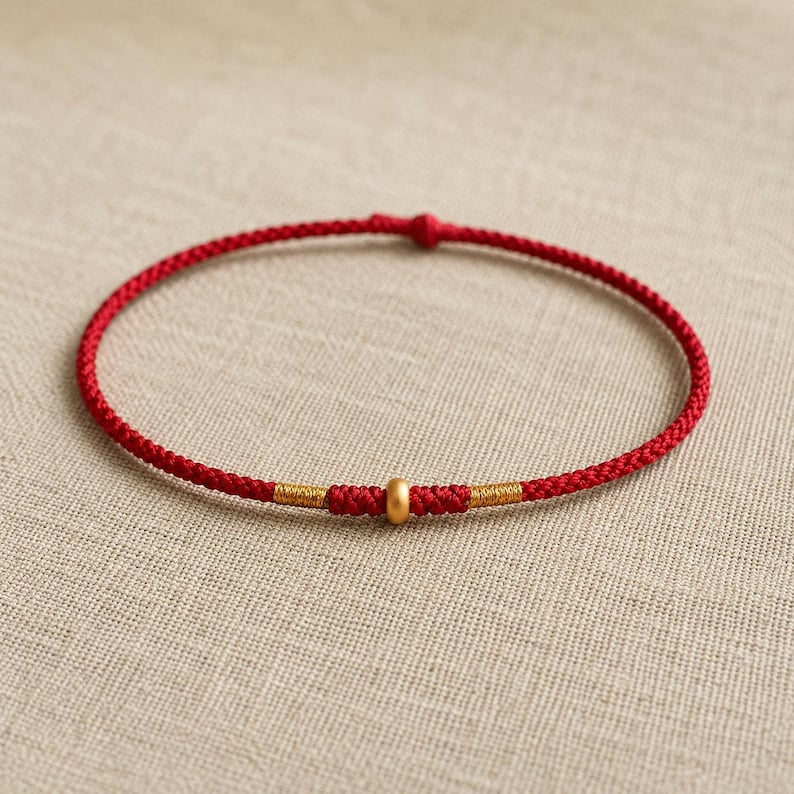 Puede incluir: Una pulsera fina de cord&oacute;n rojo con una cuenta central dorada mate, flanqueada por dos peque&ntilde;as envolturas de hilo met&aacute;lico dorado. La pulsera est&aacute; hecha de un cord&oacute;n rojo trenzado continuo con un peque&ntilde;o cierre de nudo en la parte superior.