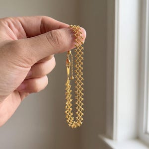 Könnte beinhalten: Ein goldfarbenes Armband mit einem filigranen, komplexen Gitter- oder Netzdesign, das aus kleinen, miteinander verbundenen Metallgliedern besteht. Das Armband verfügt über einen verstellbaren Schiebeverschluss mit einer dünnen Kette und einer kleinen, runden Endperle.