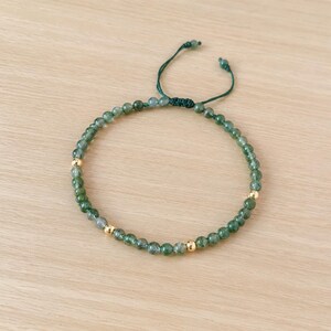 May include: A green beaded bracelet with gold-colored accents. The bracelet features small, round, green beads and three gold-colored beads. The bracelet is adjustable with a dark green cord.