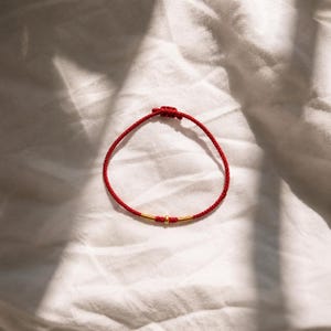 Puede incluir: Una pulsera de cord&oacute;n rojo fino que presenta una peque&ntilde;a cuenta redonda de color dorado centrada entre dos acentos cil&iacute;ndricos cortos de color dorado. La pulsera incluye un mecanismo de cierre de nudo deslizante ajustable en la parte superior.