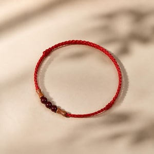 May include: A red braided bracelet with three dark red beads and two small wooden accents. The bracelet is circular and sits on a light beige surface, with shadows in the background. The bracelet is a piece of jewelry.