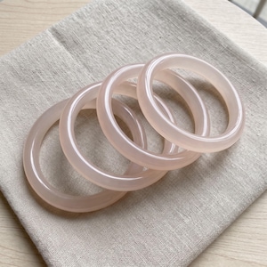 May include: A set of four smooth, translucent, pale pink stone bangles. Each bangle features a rounded, polished surface with a consistent thickness and a circular shape, showcasing a soft, uniform light pink hue throughout the material.