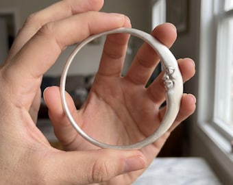 May include: A silver-toned circular bangle bracelet featuring a sculpted crescent moon face on one side. The metal has a smooth, matte finish, and the moon design includes detailed facial features such as an eye, nose, and mouth.