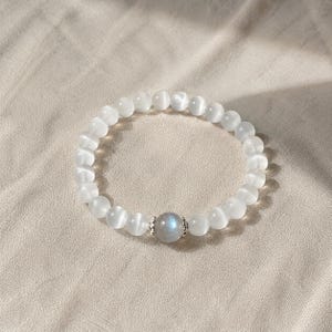 Könnte beinhalten: Ein weißes Perlenarmband mit einem zentralen, irisierenden, grau-blauen Edelstein. Das Armband besteht aus runden, undurchsichtigen weißen Perlen. Der Edelstein ist in eine silberfarbene Metalleinfassung gefasst. Das Armband wird auf einem neutralfarbenen Stoff präsentiert.