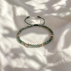 May include: Adjustable bracelet featuring small, green beads and two gold-colored beads. The bracelet is strung on a dark green cord with a sliding knot closure. The beads have a translucent quality, and the bracelet rests on a white, textured surface.