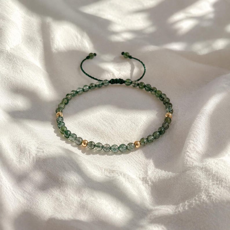 May include: Adjustable bracelet featuring small, green beads and two gold-colored beads. The bracelet is strung on a dark green cord with a sliding knot closure. The beads have a translucent quality, and the bracelet rests on a white, textured surface.