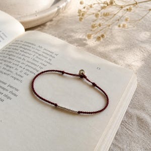 May include: A burgundy cord bracelet with a silver cylindrical bead and a small silver disc. The bracelet is resting on an open book with printed text. The background includes a white ceramic dish and a sprig of dried flowers.