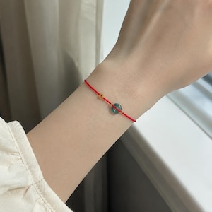 Peut inclure: Un bracelet en fil rouge avec une petite perle dorée et une pierre ronde de couleur turquoise. Le bracelet est porté au poignet.