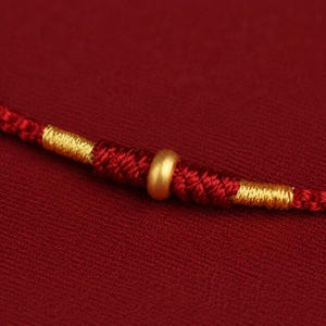 Puede incluir: Una pulsera de cord&oacute;n rojo con una cuenta dorada lisa en el centro, flanqueada por secciones de hilo rojo trenzado complejo y acentuada con envolturas de hilo dorado met&aacute;lico a ambos lados del nudo central, creando una pieza de joyer&iacute;a.