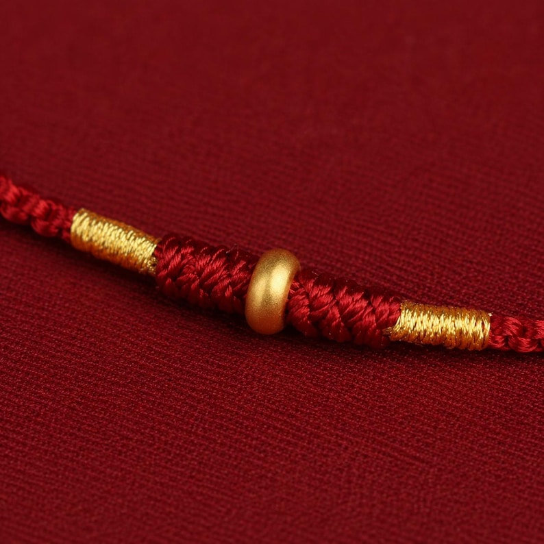 Puede incluir: Una pulsera de cord&oacute;n rojo con una cuenta dorada lisa en el centro, flanqueada por secciones de hilo rojo trenzado complejo y acentuada con envolturas de hilo dorado met&aacute;lico a ambos lados del nudo central, creando una pieza de joyer&iacute;a.