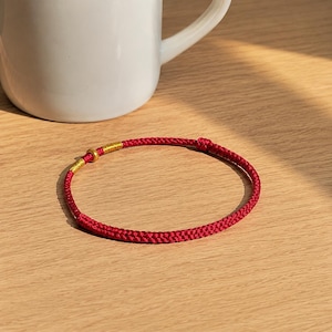 Puede incluir: Una pulsera fina de cord&oacute;n rojo con textura trenzada, acentuada con una peque&ntilde;a cuenta de metal dorado y una secci&oacute;n de hilo dorado enrollado. La pulsera tiene forma circular e incluye un peque&ntilde;o detalle de nudo a lo largo del cord&oacute;n.