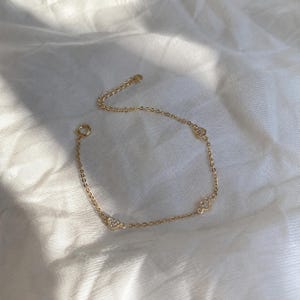 May include: A delicate gold-colored bracelet with three heart-shaped embellishments, each adorned with small, sparkling stones. The bracelet is displayed on a white, textured surface, catching the light.