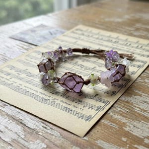 May include: A bracelet featuring raw, light purple amethyst stones encased in brown cord netting, interspersed with small, translucent white and purple flower-shaped beads and tiny green accents, arranged in a circular pattern on a flat surface.