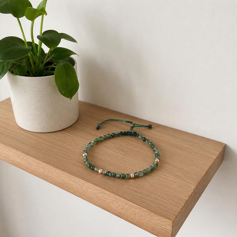 May include: A green beaded bracelet with gold-colored accents. The bracelet is on a wooden shelf next to a small potted plant with green leaves. The bracelet is adjustable with a green cord.