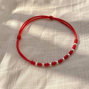 May include: A red cord bracelet featuring a series of small, polished silver-toned spherical beads interspersed with decorative knots. The bracelet is constructed from a thin, flexible red string with an adjustable sliding knot closure mechanism.